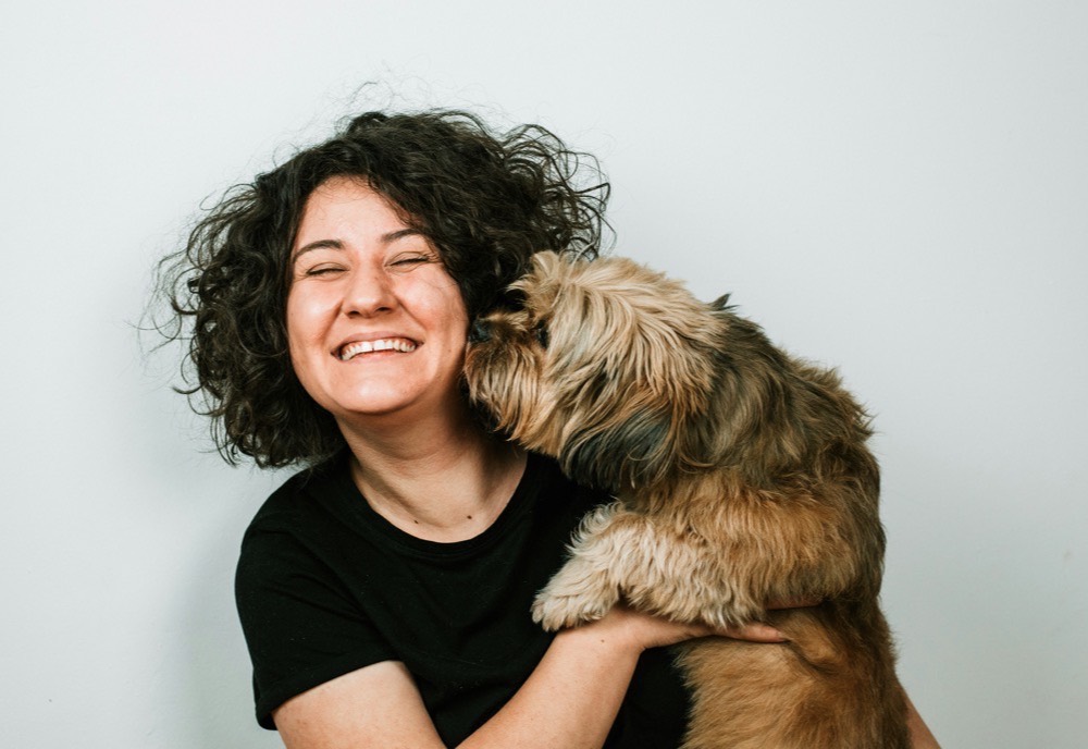A woman laughing as a small dog nuzzles her cheek.