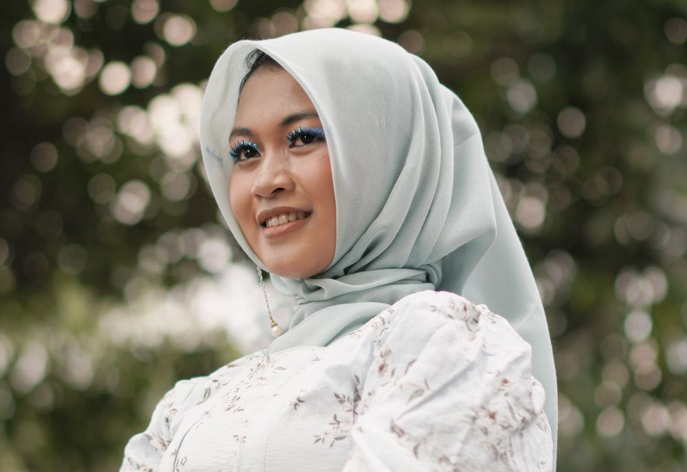 A smiling woman in a pale blue hijab and a floral blouse, outdoors.